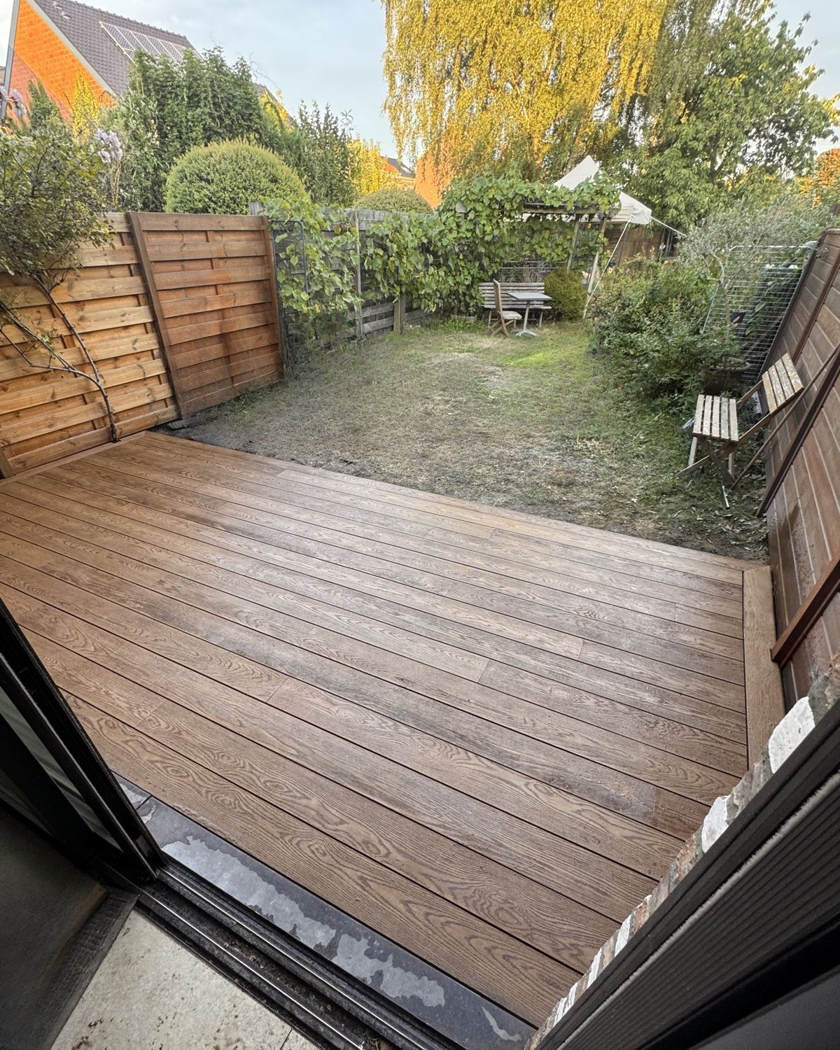 Afgewerkt terras in Millboard Enhanced Grain, kleur Coppered Oak, in de tuin, gezien vanuit het schuifraam van een bakstenen woning